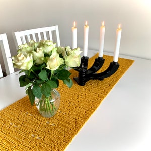May include: A yellow crocheted table runner with a black metal candle holder with four white candles on a white table with a bouquet of white roses in a clear glass vase.