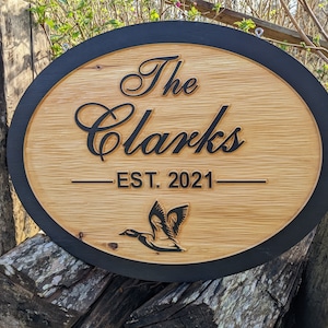 May include: Oval wooden sign with a black border. The sign says "The Clarks - EST. 2021" in black lettering. A black silhouette of a duck is in the bottom center of the sign.
