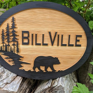 May include: Oval wooden sign with a black border. The sign has a carved design of trees, a bear, and the text "BILLVILLE".