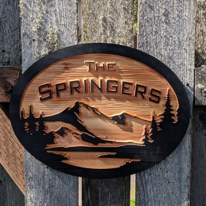 May include: A round wooden sign with a black border and a mountain scene carved into the wood. The sign says "The Springers" in black letters.