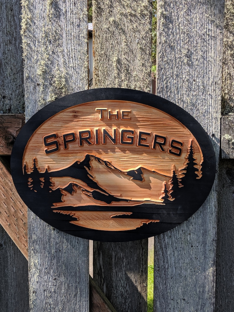 May include: A round wooden sign with a black border and a mountain scene carved into the wood. The sign says "The Springers" in black letters.