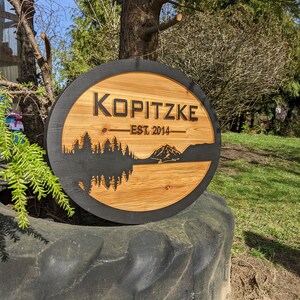 May include: A round wooden sign with a black border and a mountain scene carved into the wood. The sign says "Kopitzke Est. 2014" in black lettering.