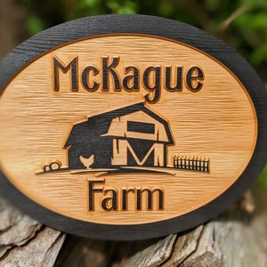 May include: A wooden oval sign with a black border. The sign says "McKague Farm" and features a black silhouette of a barn with a chicken in front of it.