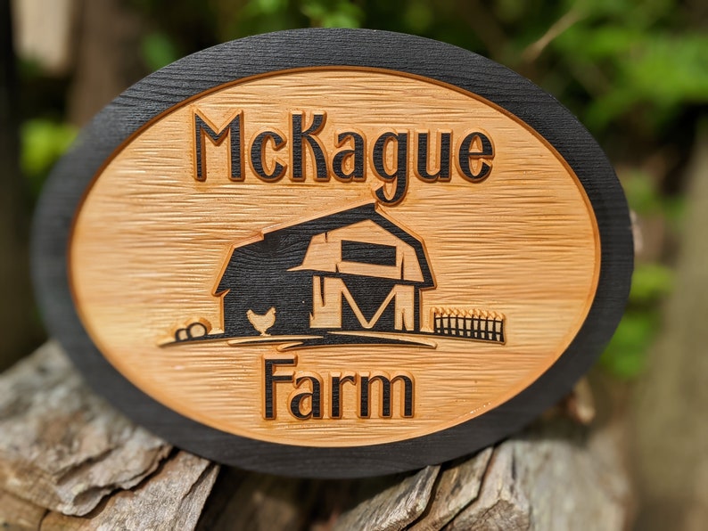 May include: A wooden oval sign with a black border. The sign says "McKague Farm" and features a black silhouette of a barn with a chicken in front of it.