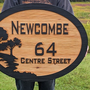 May include: A wooden oval sign with a black border. The sign has a black silhouette of a tree and a stream. The text "NEWCOMBE 64 CENTRE STREET" is carved into the wood.