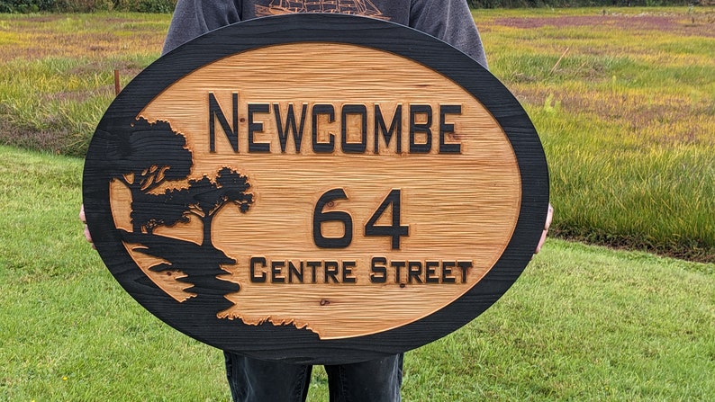 May include: A wooden oval sign with a black border. The sign has a black silhouette of a tree and a stream. The text "NEWCOMBE 64 CENTRE STREET" is carved into the wood.