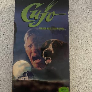 Puede incluir: Una cinta VHS de la película de terror "Cujo" basada en la novela de Stephen King. La portada muestra a una mujer gritando con un perro grande detrás de ella. El texto en la portada dice "El terror tiene un nuevo nombre..."