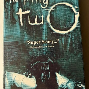 Peut inclure: Une cassette VHS du film d'horreur "The Ring Two". La couverture présente un schéma de couleurs bleu sarcelle et noir avec le titre du film et une figure fantomatique. La citation "Super Scary..." est également visible.