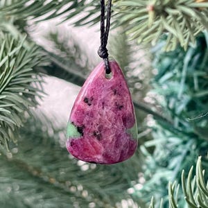 May include: A teardrop-shaped pendant with a vibrant mix of pink and green hues, suspended from a black cord. The gemstone pendant showcases a unique pattern and texture, set against a backdrop of green foliage.