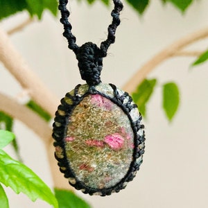 Può includere: Un ciondolo di pietra preziosa verde e rosa su una collana di cordoncino nero. Il ciondolo è di forma ovale e ha una texture naturale e ruvida.