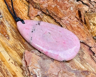 May include: A pink and white gemstone pendant with black veins, hanging from a black cord. The pendant is shaped like a teardrop and is resting on a piece of wood.