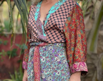 May include: A woman wearing a long, multi-coloured patterned wrap dress with a deep V-neckline. The dress has a floral print and a paisley pattern. The woman is standing in front of a green leafy background.