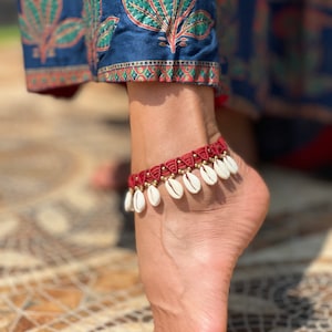 Può includere: Braccialetto da caviglia rosso e bianco con un design a conchiglia. Il braccialetto è fatto di corda e ha piccole conchiglie bianche che pendono.