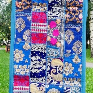 Può includere: Un arazzo colorato in patchwork con sfondo blu. L'arazzo è fatto di diversi tessuti e ha ricami intricati in filo dorato, argentato e rosa. L'arazzo è appeso a uno stendino.