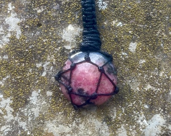 May include: A pink and gray gemstone pendant necklace. The round stone is encased in a black woven macrame net and suspended from a black cord. The background is a textured, mossy stone surface.