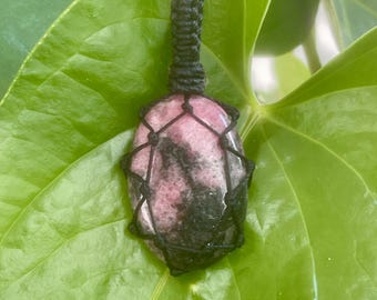 May include: A pendant necklace featuring a pink and black stone encased in a black woven macrame. The stone is oval-shaped and suspended from a black cord. The background is a large green leaf.