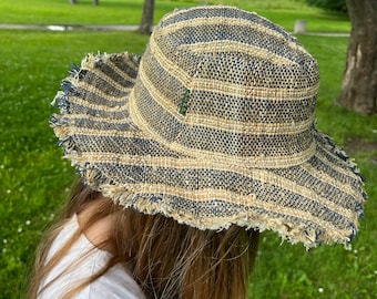 May include: A woven straw hat with a wide brim and a frayed edge. The hat features horizontal stripes in shades of beige, navy blue, and olive green. The hat is designed to provide sun protection and is suitable for outdoor activities.