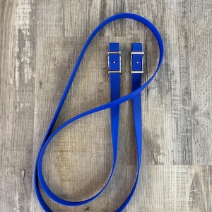 May include: A pair of bright blue stirrup leathers with silver-colored buckles. The leathers are made of a flexible material and are looped together. The background is a light brown wood grain.