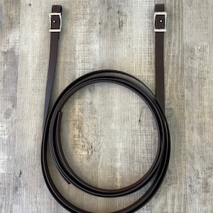 May include: Dark brown leather reins with silver-colored buckles. The reins are coiled in a circular shape, with two straps extending upwards. The background is a light gray wooden surface.