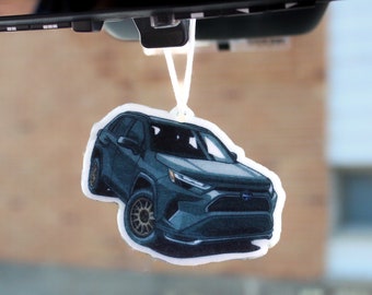 以下が含まれることがあります： 車のバックミラーから吊り下げられた、濃い青色のSUVの形をした芳香剤。 車両はブロンズ色のホイールを備え、詳細なイラストスタイルで描かれています。 背景はぼやけており、車のインテリアを示唆しています。
