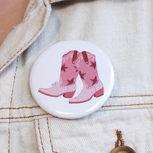 May include: A white button pin featuring a pair of pink cowboy boots with star accents. The pin is pinned to a light-colored denim jacket. The boots have a western theme.