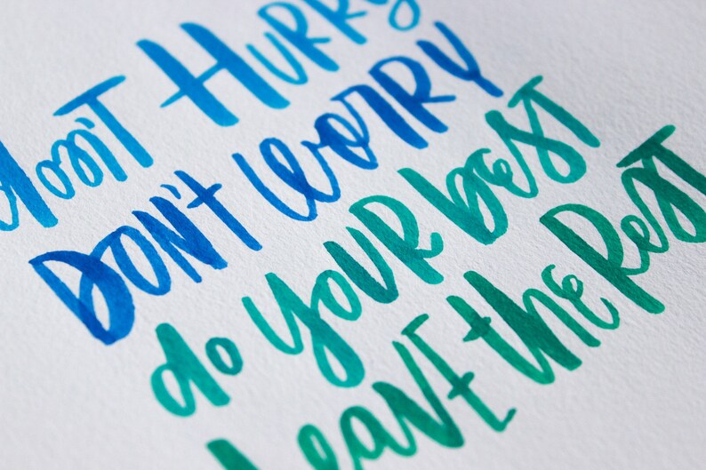 May include: Hand-lettered calligraphy in blue and green shades on white paper. The text reads "Don't hurry, don't worry, do your best, leave the rest."