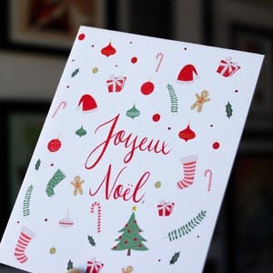 K&ouml;nnte beinhalten: Eine wei&szlig;e Weihnachtskarte mit roten und gr&uuml;nen Illustrationen von Weihnachtsornamenten, Zuckerstangen, einem Lebkuchenmann, einem Strumpf, einem Weihnachtsbaum und einer Weihnachtsm&uuml;tze. Die Karte sagt "Joyeux Noel" in roter Kursivschrift.