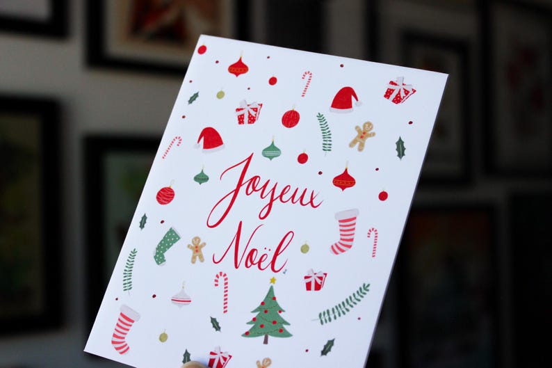 K&ouml;nnte beinhalten: Eine wei&szlig;e Weihnachtskarte mit roten und gr&uuml;nen Illustrationen von Weihnachtsornamenten, Zuckerstangen, einem Lebkuchenmann, einem Strumpf, einem Weihnachtsbaum und einer Weihnachtsm&uuml;tze. Die Karte sagt "Joyeux Noel" in roter Kursivschrift.