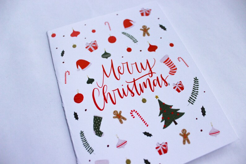 K&ouml;nnte beinhalten: Eine wei&szlig;e Weihnachtskarte mit einer roten "Frohe Weihnachten"-Botschaft, umgeben von festlichen Illustrationen von Ornamenten, Zuckerstangen, Lebkuchenm&auml;nnern, Str&uuml;mpfen und einem Weihnachtsbaum.