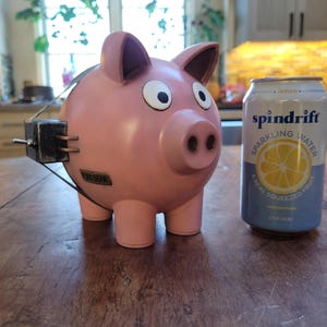 May include: A pink piggy bank with googly eyes and a metal band around its middle. The piggy bank is sitting on a wooden table next to a can of Spindrift sparkling water with a lemon slice on the label.
