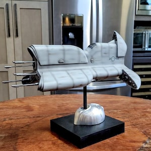 Puede incluir: Maqueta gris de una nave espacial con base negra y soporte plateado. La nave espacial tiene múltiples aletas y un diseño detallado. El modelo se exhibe sobre una mesa de madera.