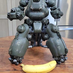 Pode incluir: Modelo detalhado verde-oliva de um robô bípede com múltiplos acessórios de armas. Uma banana amarela é colocada em frente ao robô para escala. O robô está em uma superfície de madeira.