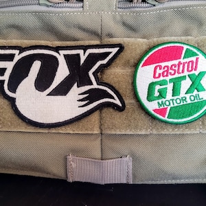 Puede incluir: Dos parches cosidos en un chaleco táctico de color beige. El parche de la izquierda es blanco y negro y dice "FOX" con una cola de zorro estilizada. El parche de la derecha es un círculo rojo, blanco y verde con el texto "Castrol GTX MOTOR OIL".