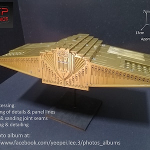 Puede incluir: Un modelo de nave espacial de color dorado con detalles intrincados, que mide aproximadamente 33 cm de largo, 18 cm de ancho y 76 cm de alto. El modelo está sobre un soporte negro y tiene un diseño detallado con un emblema en forma de escudo en la parte delantera.
