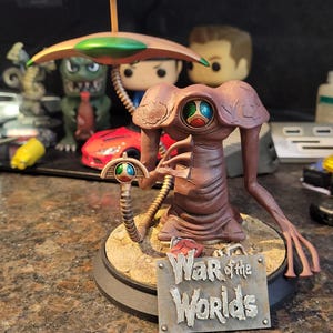 May include: A brown War of the Worlds alien figurine with a large head, long arms, and a ray gun. The figurine has a green and red eye and is standing on a circular base with the words "War of the Worlds" on a sign.