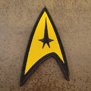 Op de afbeelding: Geborduurde Star Trek-insignia-patch in geel en zwart. De patch heeft een gele pijlpuntvorm met een zwarte ster en rand. Ontworpen om op stof te naaien of te strijken.