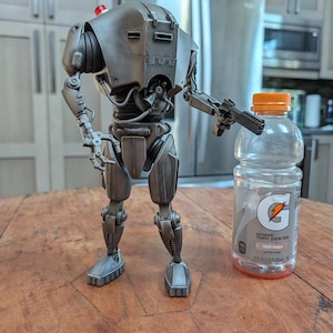 May include: A detailed, metallic gray robot figure with a gun, standing next to a 28 fl oz Gatorade bottle. The robot has a red light on its head and intricate mechanical details. The bottle is clear with an orange cap and a Gatorade logo.