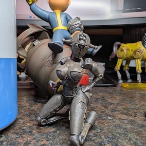 May include: A detailed image of Fallout-themed figurines. A gray robot figure kneels in the foreground. Behind it, a Vault Boy figurine sits atop a miniature bomb. A yellow horse figurine is visible in the background.