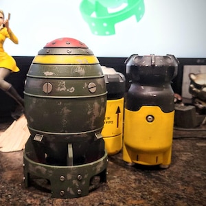 May include: A collection of detailed, weathered, model bombs with a red, yellow, and green color scheme. The models have a vintage aesthetic. A figurine of a woman in a yellow dress is in the background.
