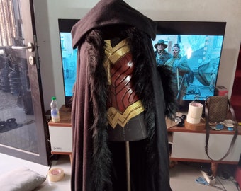 May include: A black hooded cloak with a faux fur trim. The cloak is worn over a red and gold costume with a sculpted bodice.