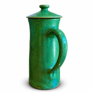 May include: A tall, green ceramic pitcher with a matching lid. The pitcher has a large, curved handle and a slightly textured surface. The color is a vibrant, solid green, and the pitcher appears to be handmade. The lid has a small knob on top.
