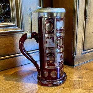 May include: A red glass beer stein with a pewter lid and a handle. The stein is decorated with etched images of buildings and the words "Bollene" and "Villars".