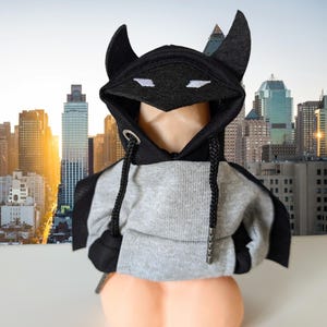 May include: A gray and black hooded sweatshirt with a black bat-like hood and ears. The sweatshirt has a drawstring and a zipper. The background is a cityscape.