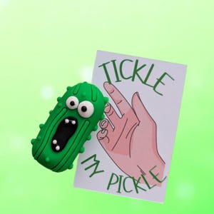 May include: A green pickle-shaped figurine with white eyes and an open mouth, next to a card with a hand illustration and the text "TICKLE MY PICKLE" in green. The background is a gradient of light green.