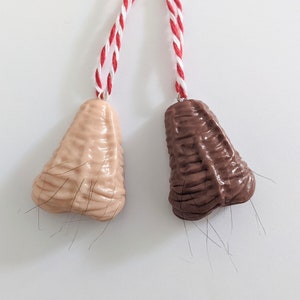May include: Two nose-shaped ornaments with brown and beige hair. The ornaments are hanging from red and white strings.