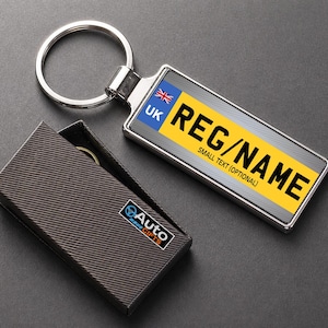 May include: A silver rectangular keyring with a UK number plate design. The plate has the text "REG/NAME" in black on a yellow background. The keyring is attached to a silver ring. A black gift box with the "Auto Gifts" logo is next to the keyring.