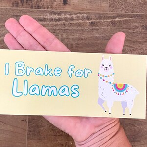 May include: A yellow bumper sticker with the text "I Brake for Llamas" in blue lettering. A cartoon llama with a colourful saddle and beaded necklace is on the right side of the sticker.