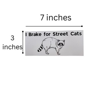 May include: A white sticker with black text and a black and white illustration of a raccoon. The text reads "I Brake for Street Cats". The sticker is 17.8 cm wide and 7.6 cm tall.
