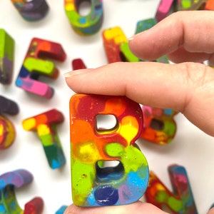 May include: A colorful crayon letter "B" held between fingers, with other rainbow-colored letters in the background. The letter is a mix of red, orange, yellow, green, and blue. The crayons appear handmade and are on a white surface.