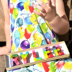 May include: A child wearing a sleeveless jumpsuit with a colorful balloon print holds a tray of rainbow-colored crayons spelling out the name "RYLEE". The crayons are in a rectangular box.